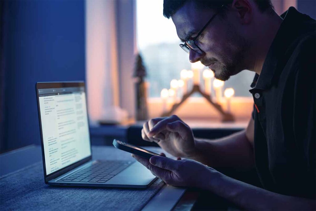 Person using a smartphone while working on a laptop in a dimly lit room